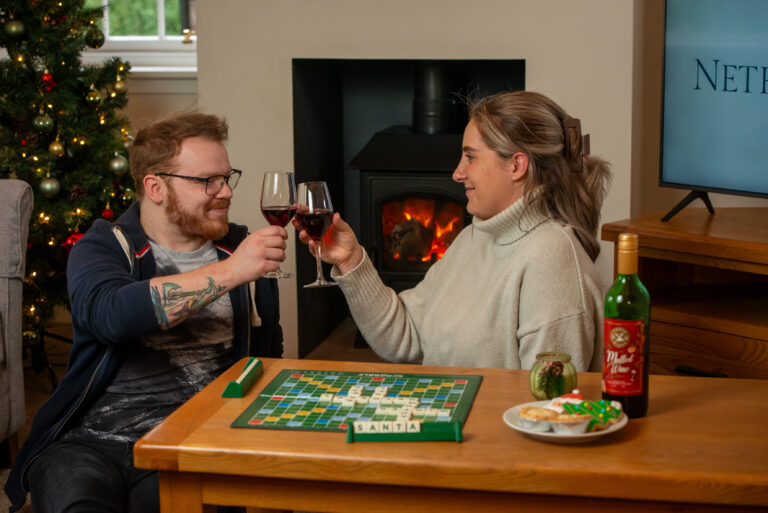 A cosy scene inside a Netherby Hall self-catering apartment. Two people sit at a table playing Scrabble by a lit fireplace, raising glasses of wine in a toast. A Christmas tree with festive decorations is visible in the background. The table is set with snacks, including biscuits, and a bottle of mulled wine. The atmosphere is warm and celebratory, perfect for a winter holiday.