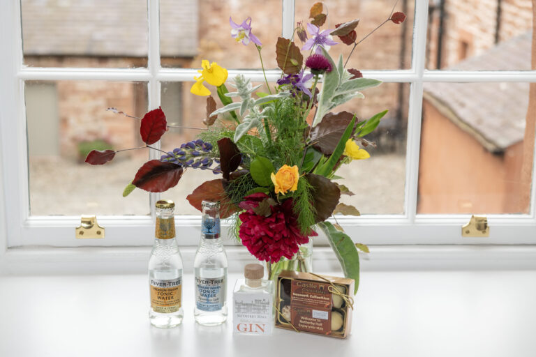 A beautifully arranged bouquet of fresh flowers sits on a windowsill next to bottles of Fever-Tree tonic water and a bottle of Netherby Hall gin, accompanied by a box of Castle Chocolates, creating a welcoming atmosphere for guests at the luxury self-catering accommodation in Cumbria.