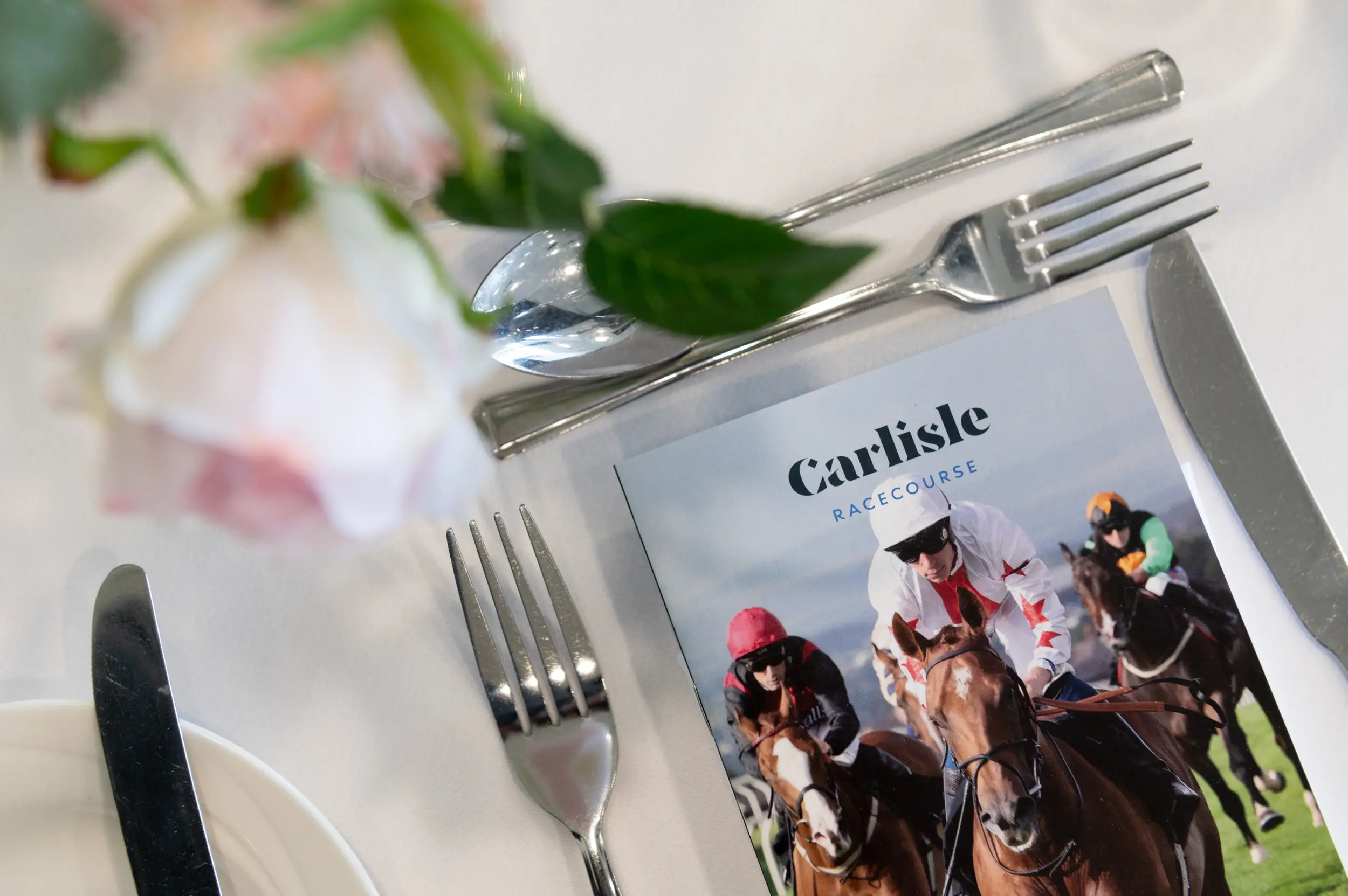 A close-up image of a dining table setting featuring a menu for Carlisle Racecourse, surrounded by cutlery including a knife, fork, and spoon, with a blurred floral arrangement in the foreground.