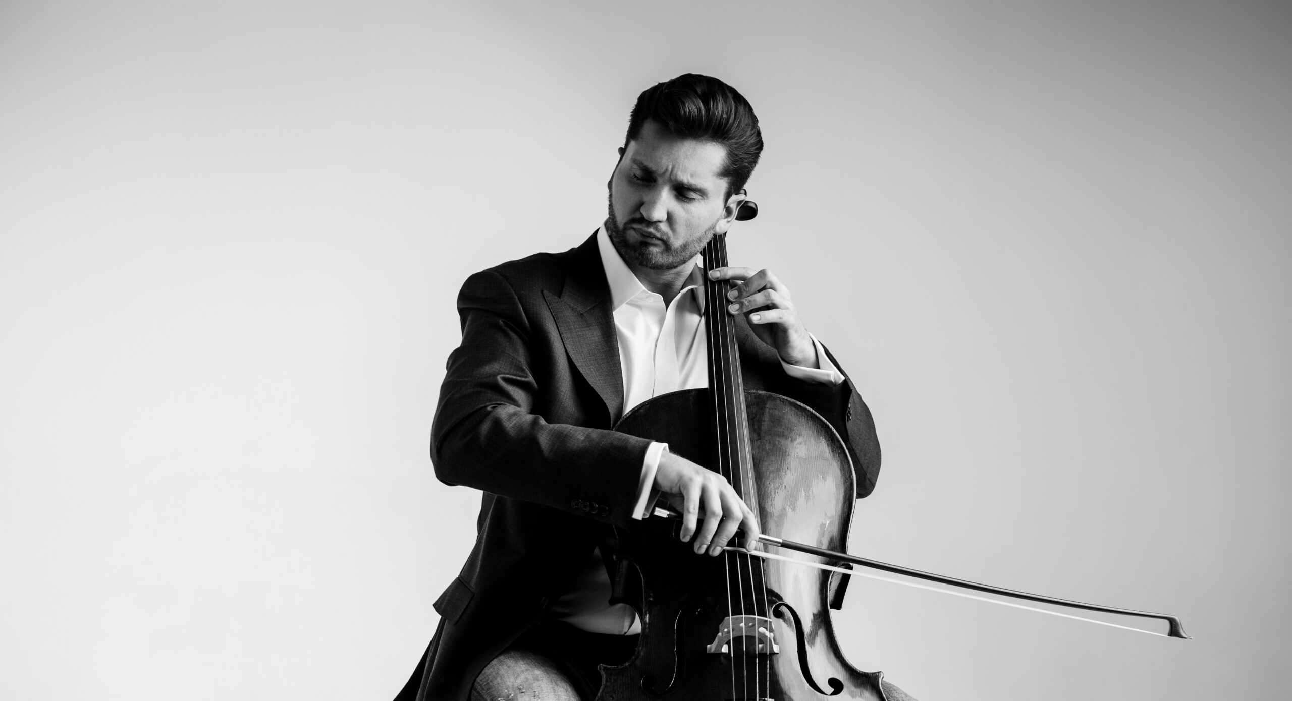 A man in a suit playing a cello, showcasing musical talent in a black and white studio setting.