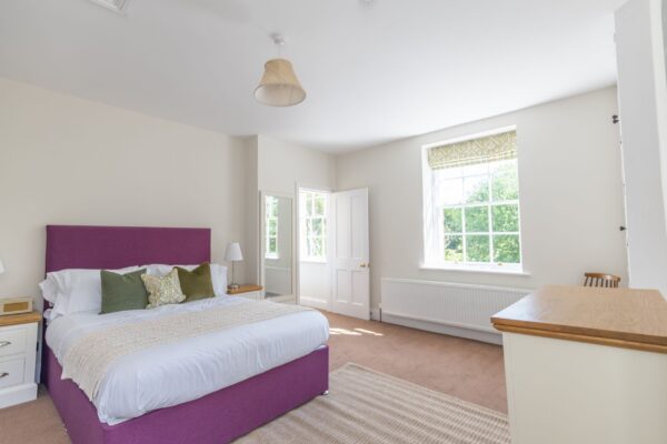 Brightly lit bedroom in the Coach House apartment featuring a purple bed frame, white linens, and large window at Netherby Hall, Cumbria