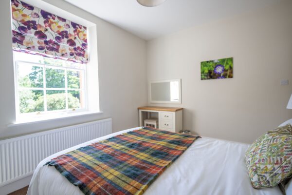 Bedroom at the Coach House apartment featuring a colorful patterned Roman blind, a plaid throw on the bed, and a vanity mirror
