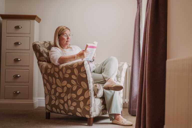 A woman relaxing and reading a book in a patterned armchair inside the luxury Engineer Apartment at Netherby Hall