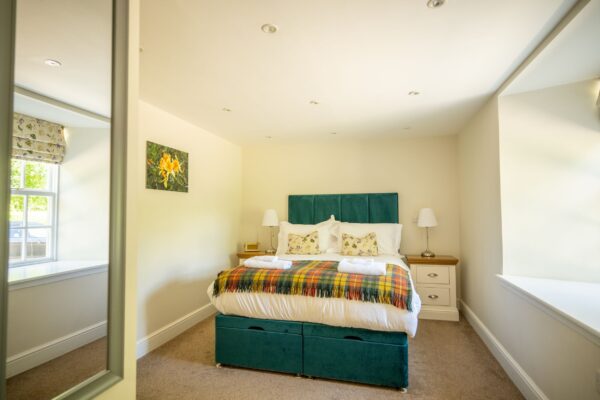 Bright bedroom at Gardener's Cottage featuring a bed with a teal velvet headboard and colorful plaid throw at Netherby Hall, Cumbria
