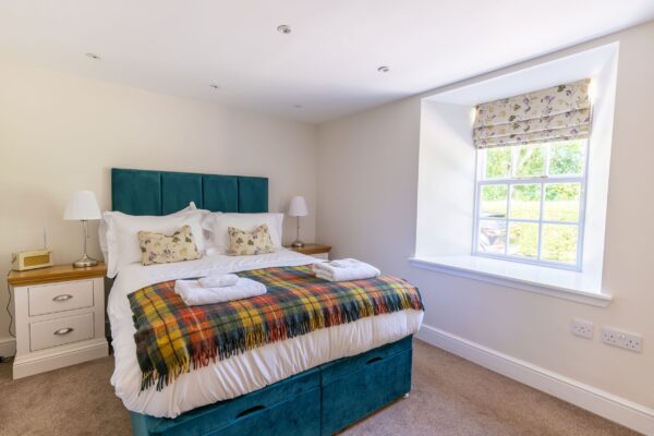 Bedroom in the Gardener's Cottage featuring a teal velvet bed, patterned throw, and large window at Netherby Hall, Cumbria