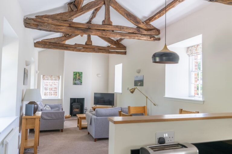 Living room in Gardener's Cottage featuring rustic exposed wooden beams, comfortable grey sofas, and a cozy fireplace