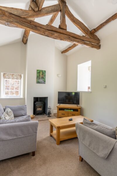 Living room in Gardener's Cottage featuring rustic wooden ceiling beams, a grey sofa, wooden coffee table, and a black wood-burning stove