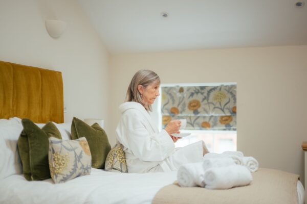 Woman in a white bathrobe relaxing on a bed in the luxury Grooms' Quarters apartment at Netherby Hall, Cumbria