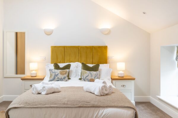 Modern bedroom in Grooms' Quarters apartment at Netherby Hall, featuring a mustard velvet headboard, white linens, and cozy bedside lamps
