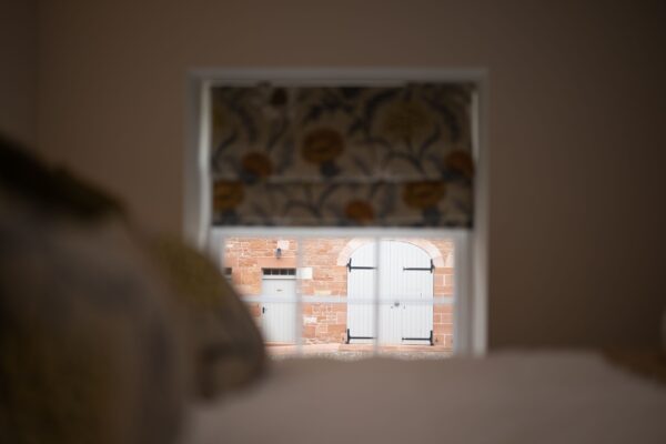 View through a window at Grooms' Quarters apartment showing stone buildings and a white door at Netherby Hall, Cumbria