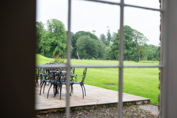 View through a window of the patio and lush green garden at Grooms' Quarters, luxury self-catering accommodation at Netherby Hall, Cumbria