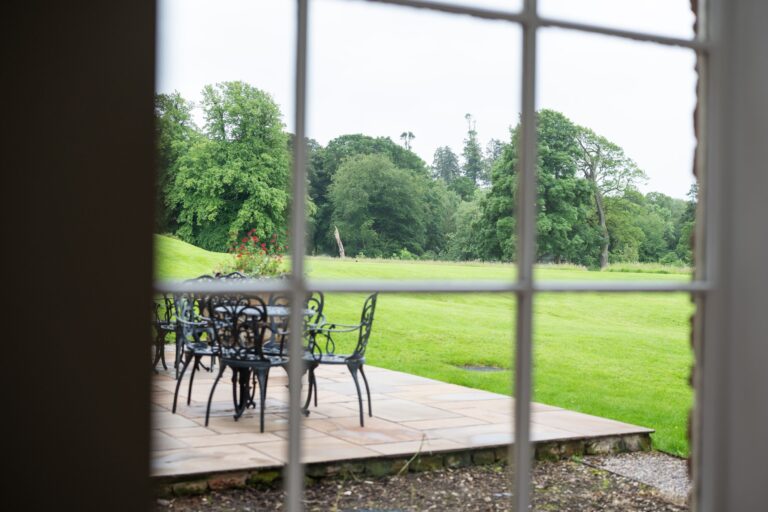 View through a window of the patio and lush green garden at Grooms' Quarters, luxury self-catering accommodation at Netherby Hall, Cumbria