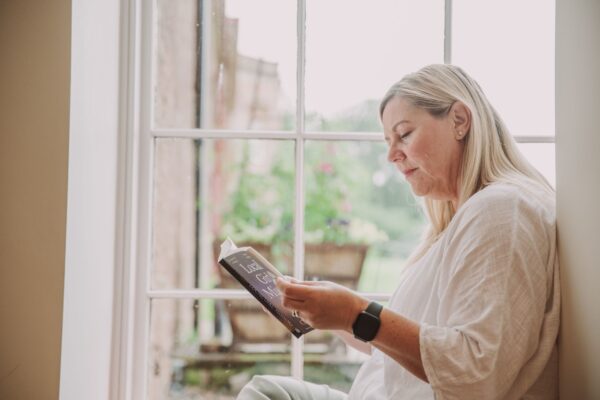 Woman reading a book by a large window in a luxury Grooms' Quarters apartment at Netherby Hall, Cumbria