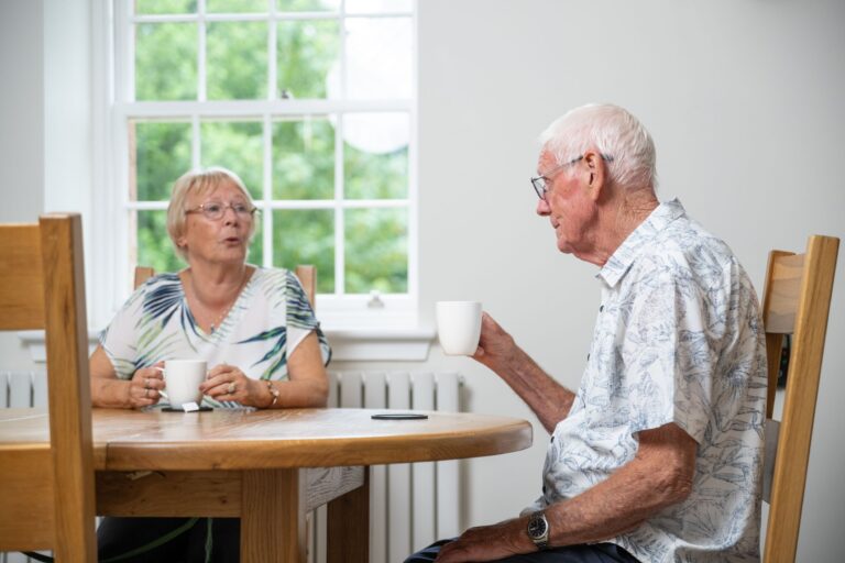 An elderly couple enjoys coffee and conversation while sitting at a wooden table inside a bright Independent Apartment at Netherby Hall