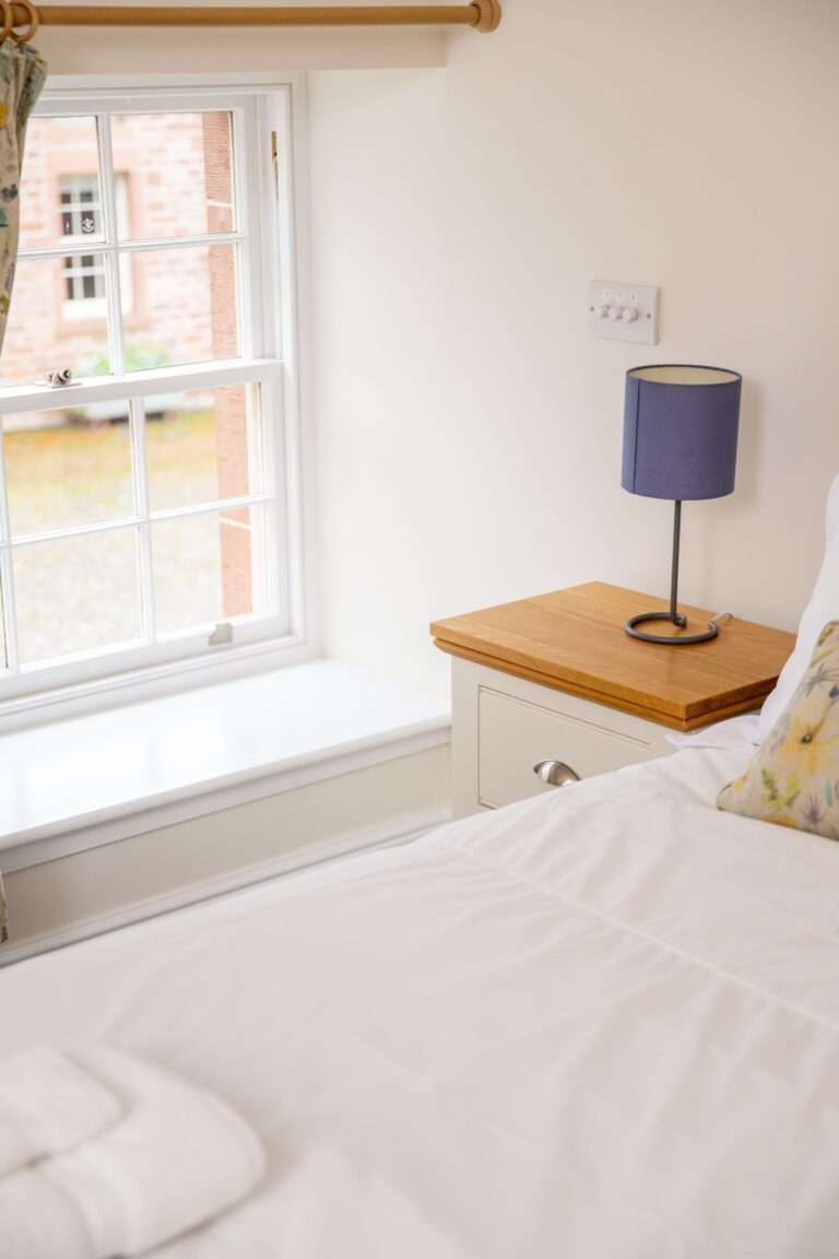 Bedroom in Independent Apartment at Netherby Hall featuring a white bed, wooden nightstand with a purple lamp, and a large window