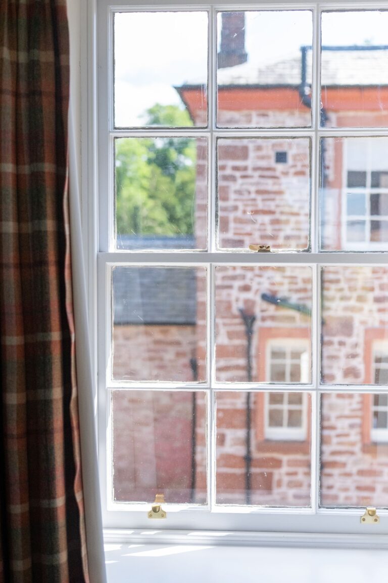 View through a multi-pane window in the Salutation Apartment at Netherby Hall, showing historic stone buildings in Cumbria