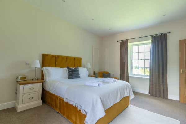 Brightly lit bedroom in the luxury Salutation Apartment at Netherby Hall featuring a large bed with a mustard velvet headboard and neutral tones