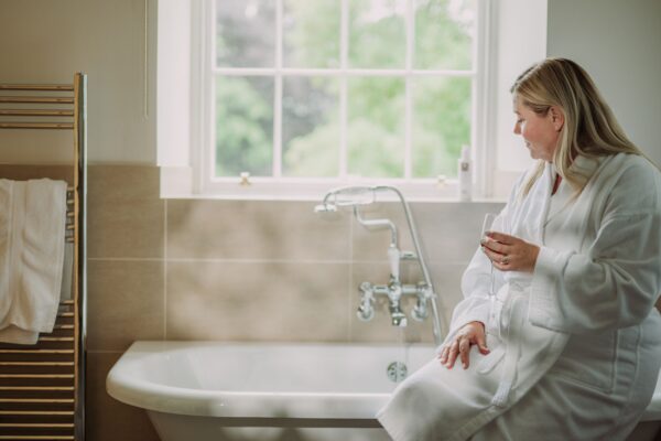 Woman in a white robe relaxing with a drink by a bathtub in the luxury Sir Walter Scott Apartment at Netherby Hall