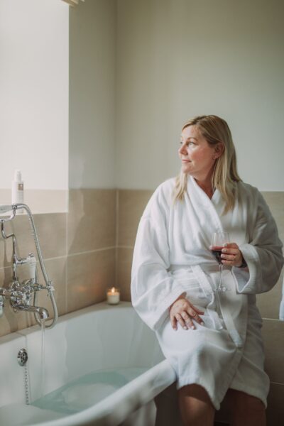 A woman in a white bathrobe holding a glass of wine, relaxing in the luxury Sir Walter Scott Apartment at Netherby Hall, Cumbria