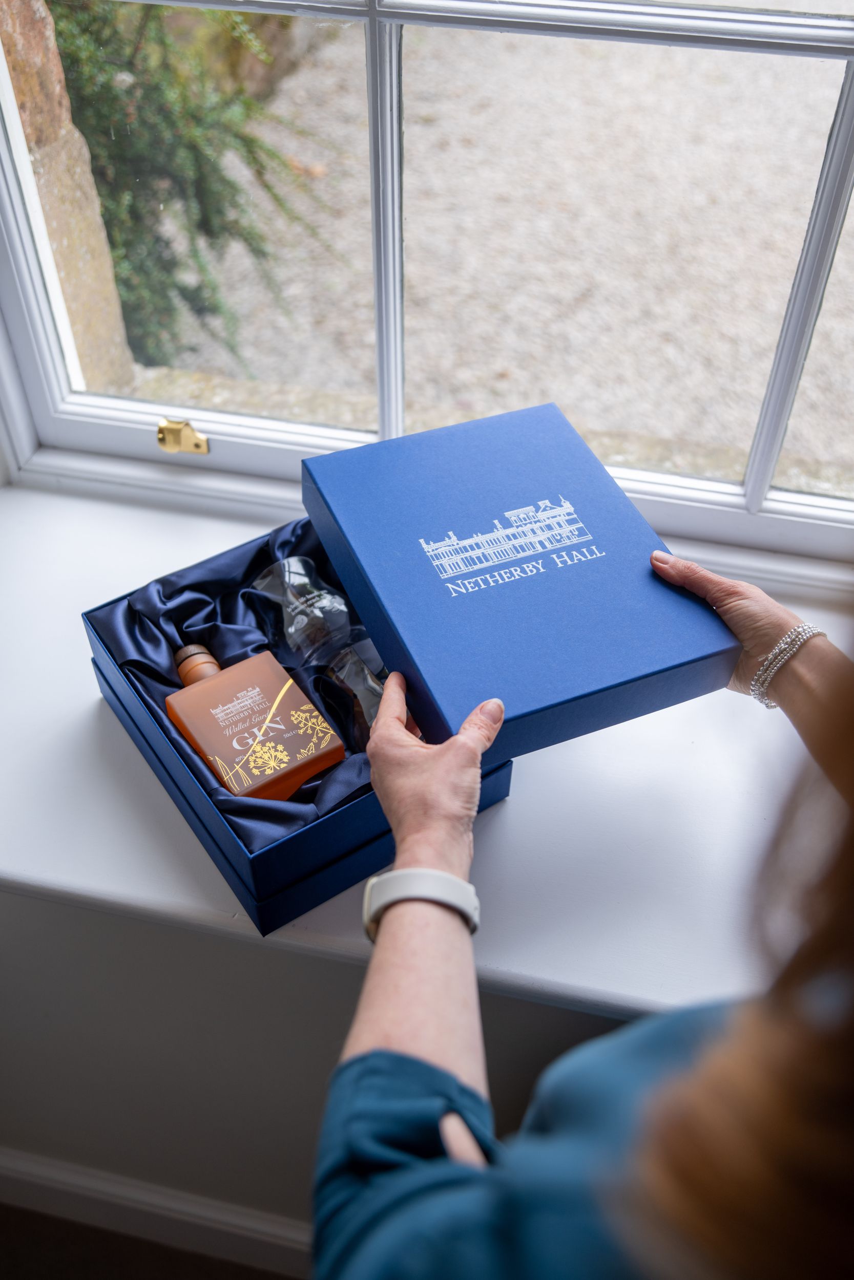 Netherby Hall gin bottle and blue gift box with glasses set against a lush garden background A person opening a luxury blue welcome gift box containing gin and glassware at Netherby Hall, Cumbria