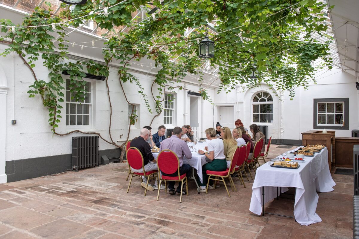Guests dine on red chairs in the ivy-covered conservatory at Netherby Hall with a table of snacks nearby