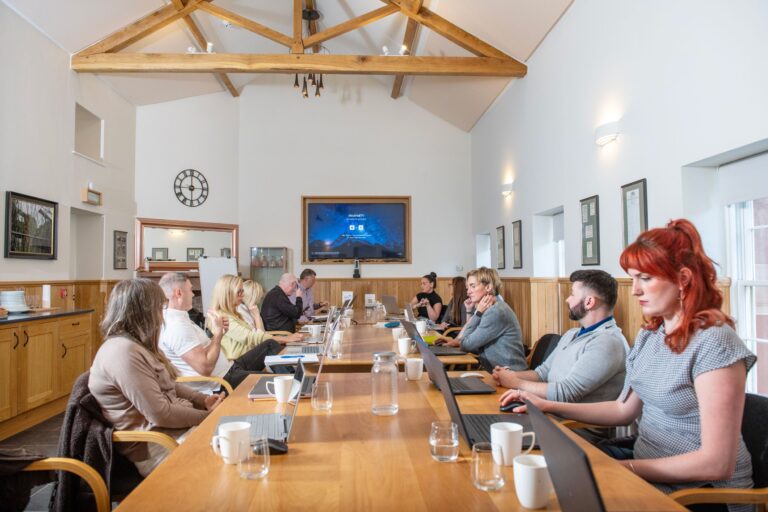 Attendees at a Netherby Hall boardroom meeting seated at a long table with laptops facing a large display screen