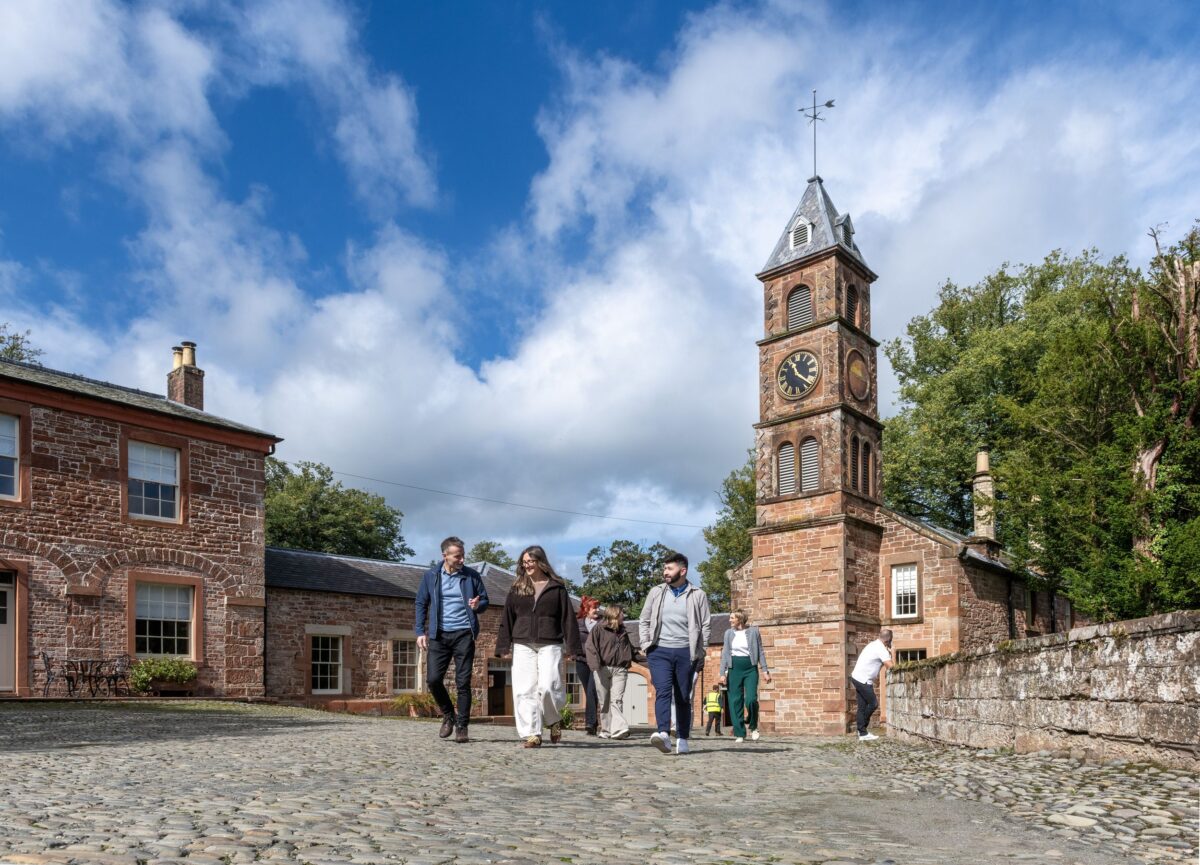 Visitors stroll across a cobblestone courtyard at Netherby Hall with the historic clock tower and sandstone buildings under a blue sky