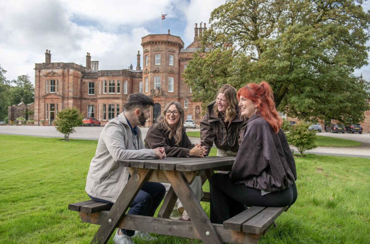 Four friends laughing at a wooden picnic table on the lawn with Netherby Hall manor house in the background