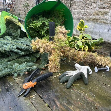 Festive foliage craft: moss cross base, evergreen cuttings, secateurs and gloves on a wooden table at Netherby Hall.