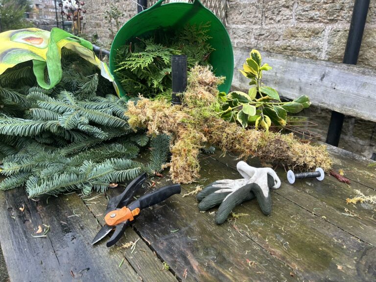 Festive foliage craft: moss cross base, evergreen cuttings, secateurs and gloves on a wooden table at Netherby Hall.